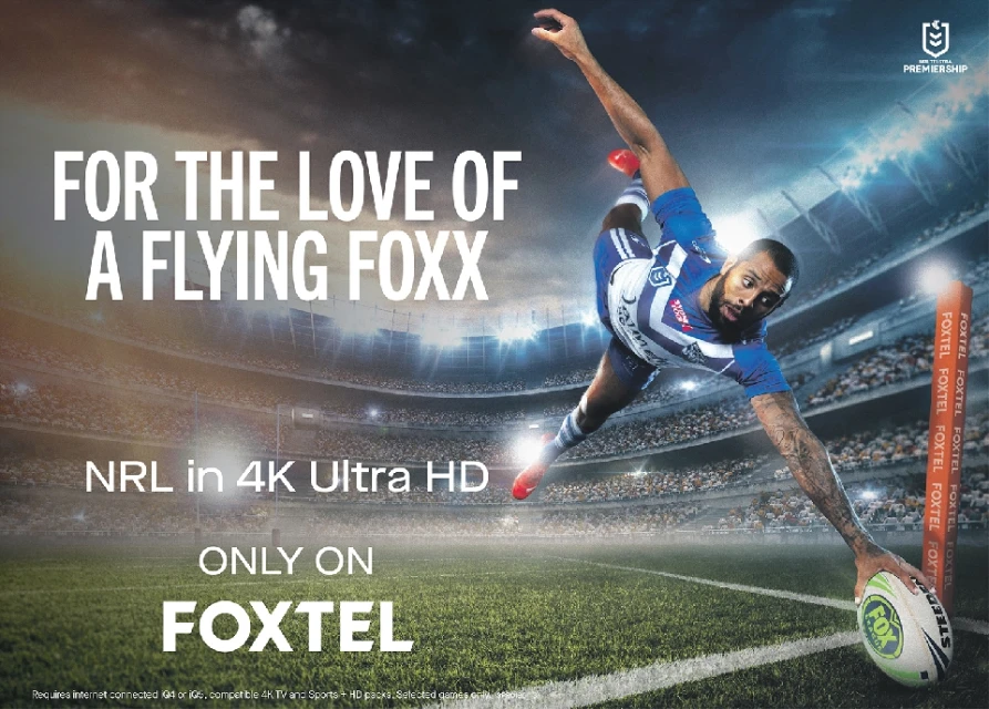 A football player stretches his arm towards a ball in a stadium with a Foxtel advertisement featuring text: 'For the Love of a Flying Foxx'.