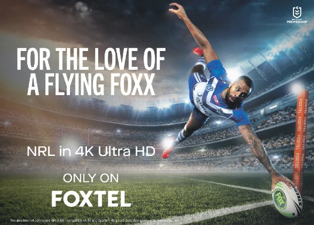 A football player stretches his arm towards a ball in a stadium with a Foxtel advertisement featuring text: 'For the Love of a Flying Foxx'.