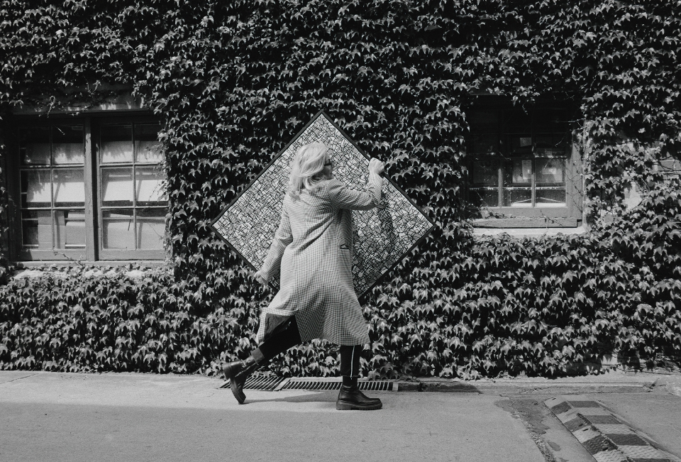Woman in patterned coat walks past ivy-covered building holding framed art. Black and white photography, geometric patterns, urban outdoor scene.