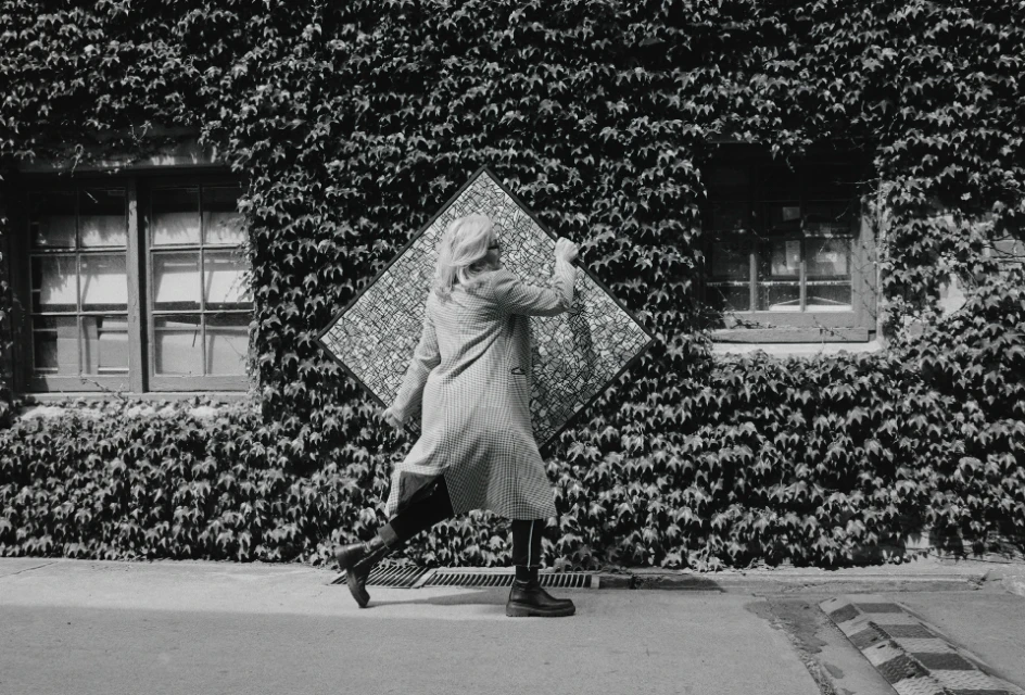 Woman in patterned coat walks past ivy-covered building holding framed art. Black and white photography, geometric patterns, urban outdoor scene.