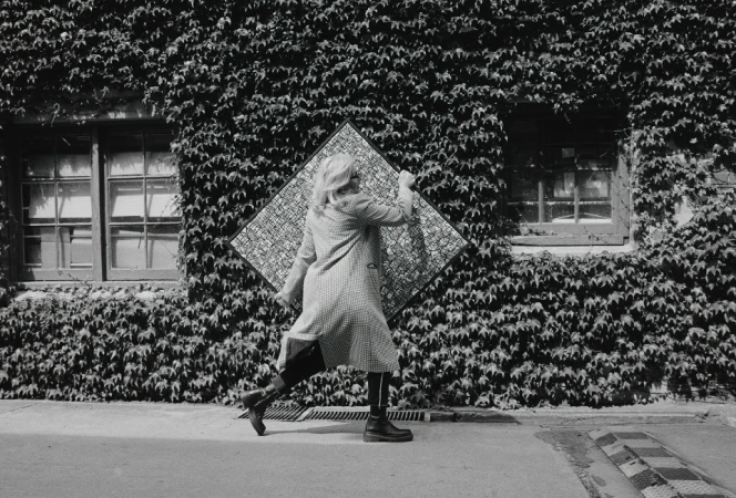 Woman in patterned coat walks past ivy-covered building holding framed art. Black and white photography, geometric patterns, urban outdoor scene.