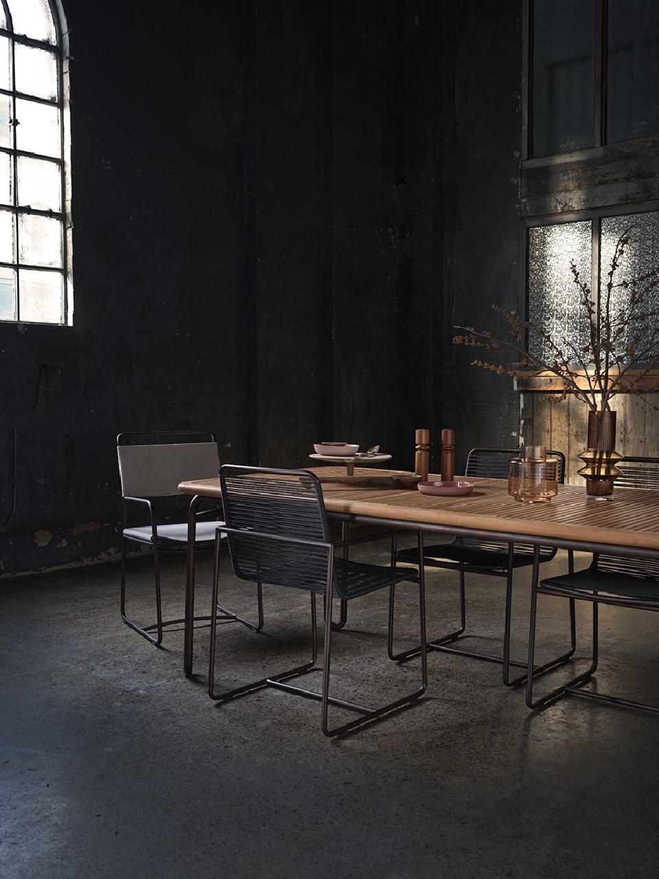 Contemporary dining room with a light wood table surrounded by metal chairs, set against a dark, moody interior and natural light.