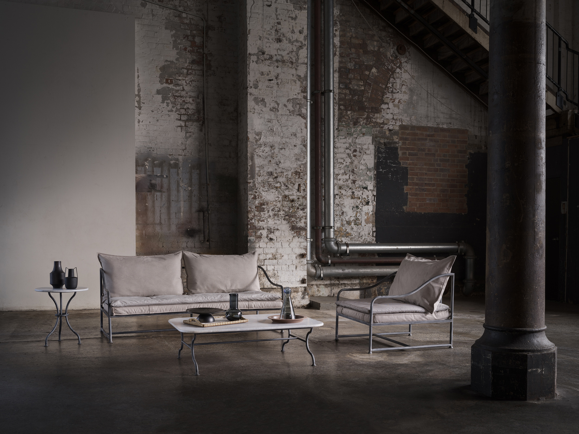 Living room interior with sofa, armchair, marble table, decorative objects, and industrial-style wall in a neutral color palette.
