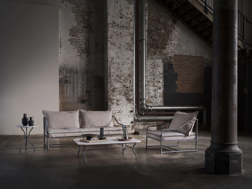 Living room interior with sofa, armchair, marble table, decorative objects, and industrial-style wall in a neutral color palette.
