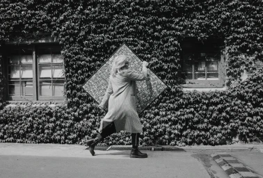 Woman in patterned trench coat carrying a large diamond-shaped framed artwork past an ivy-covered building exterior.