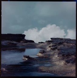 Ocean waves crashing against dark rocky coastal shelf with a tide pool under an overcast sky by Grant Harvey