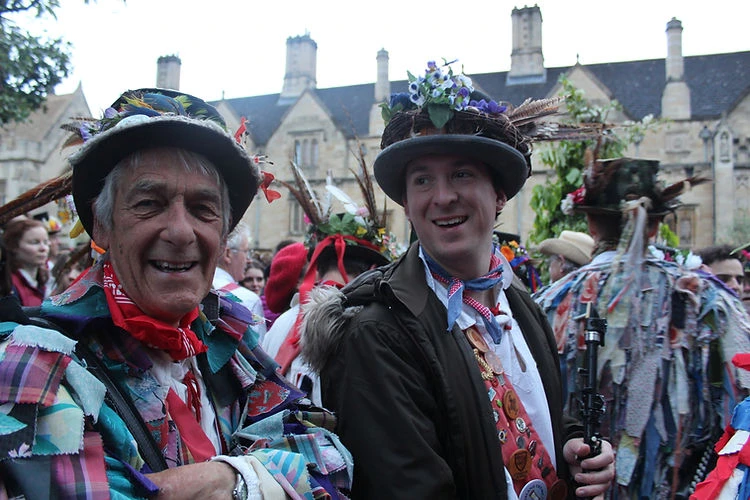 Two adult males smiling in traditional morris dancing costumes with hats and colorful patchwork jackets outdoors near stone buildings.