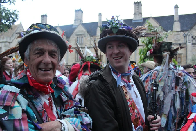 Two adult males smiling in traditional morris dancing costumes with hats and colorful patchwork jackets outdoors near stone buildings.