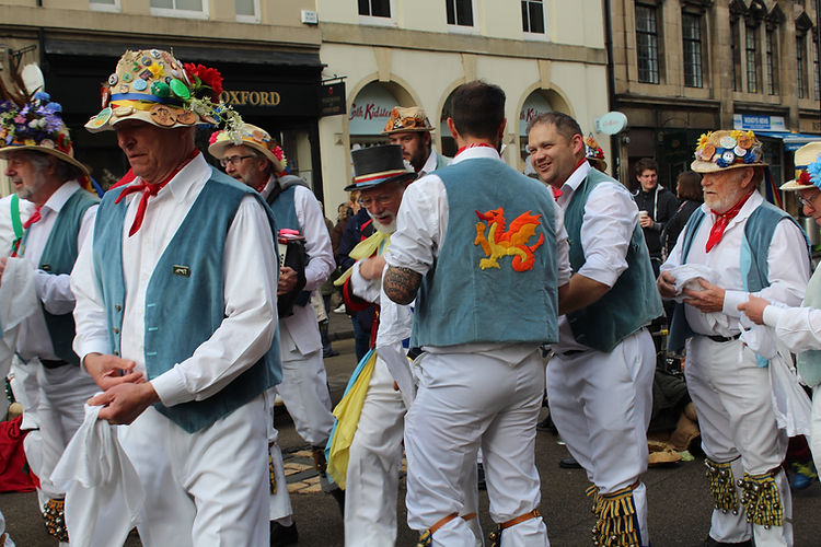 Group of Morris dancers performing outdoors, wearing traditional costumes with vests, hats, white pants, and bells on their ankles.