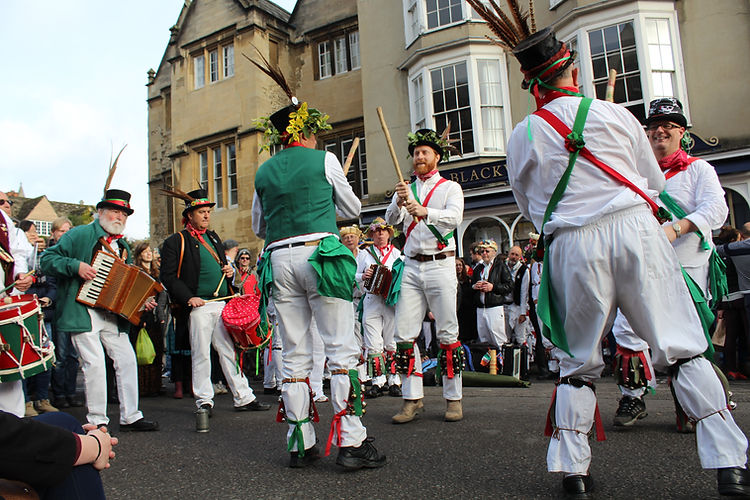 Outdoor performance featuring a group of Morris dancers in traditional attire, including hats, bells, and ribbons, performing on the street.