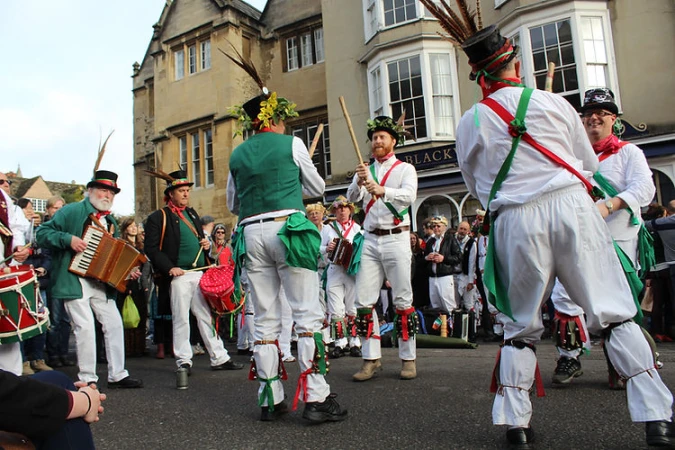 Outdoor performance featuring a group of Morris dancers in traditional attire, including hats, bells, and ribbons, performing on the street.