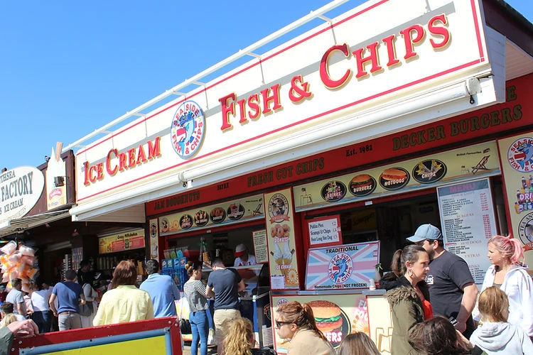 Outdoor daytime shot of a seaside food stand serving fish and chips, ice cream, and burgers, with a crowd of people in front.