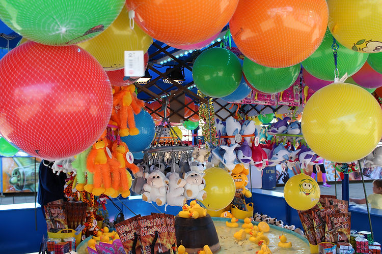 Colorful balls and stuffed animals hang above a game booth with yellow rubber ducks at an outdoor fair or carnival attraction.
