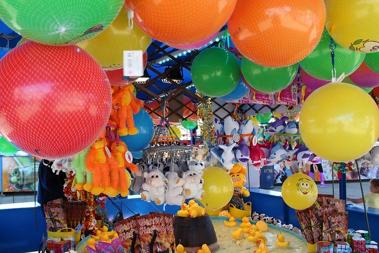 Colorful balls and stuffed animals hang above a game booth with yellow rubber ducks at an outdoor fair or carnival attraction.