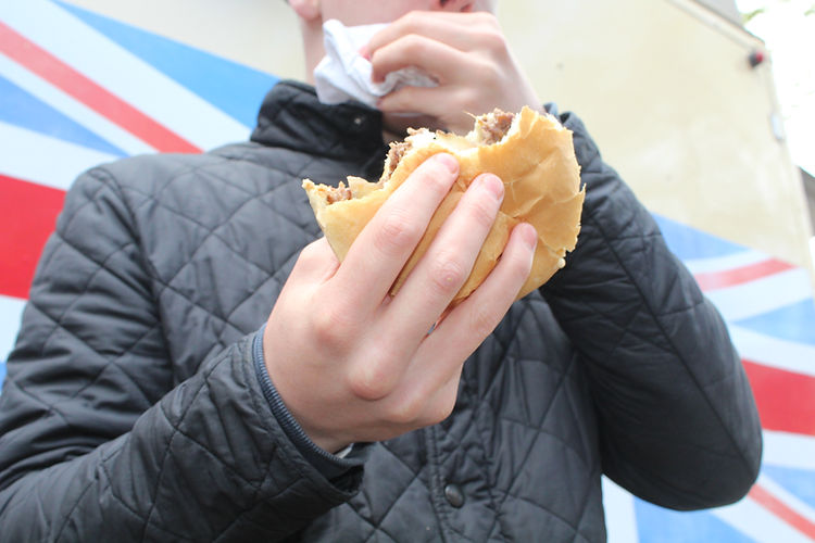 Close-up of a young person holding a half-eaten burger with both hands, outdoors, wearing a black quilted jacket, with a patterned background.