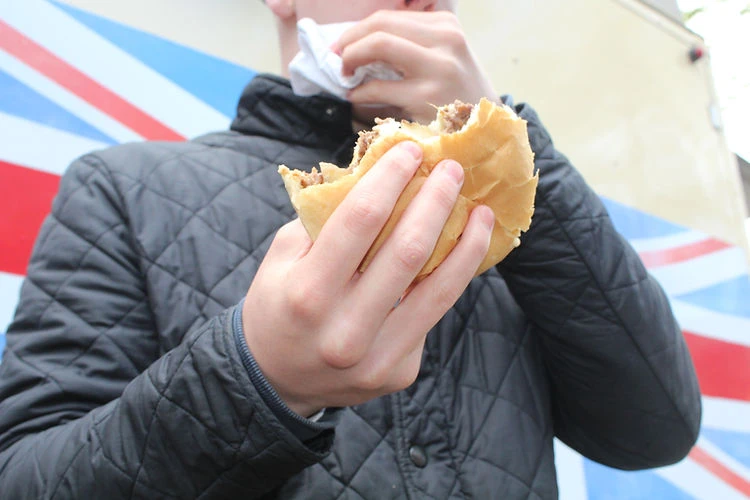 Close-up of a young person holding a half-eaten burger with both hands, outdoors, wearing a black quilted jacket, with a patterned background.