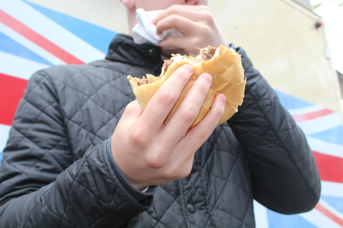 Close-up of a young person holding a half-eaten burger with both hands, outdoors, wearing a black quilted jacket, with a patterned background.