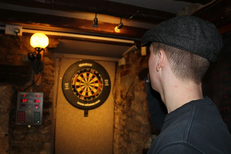 Adult male wearing a cap in a dark room standing next to a dartboard and electronic scoring system mounted on a stone wall.