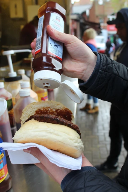 Person squeezes a bottle of barbecue sauce onto a sausage in a bun held with a paper napkin, taken outside in daylight.