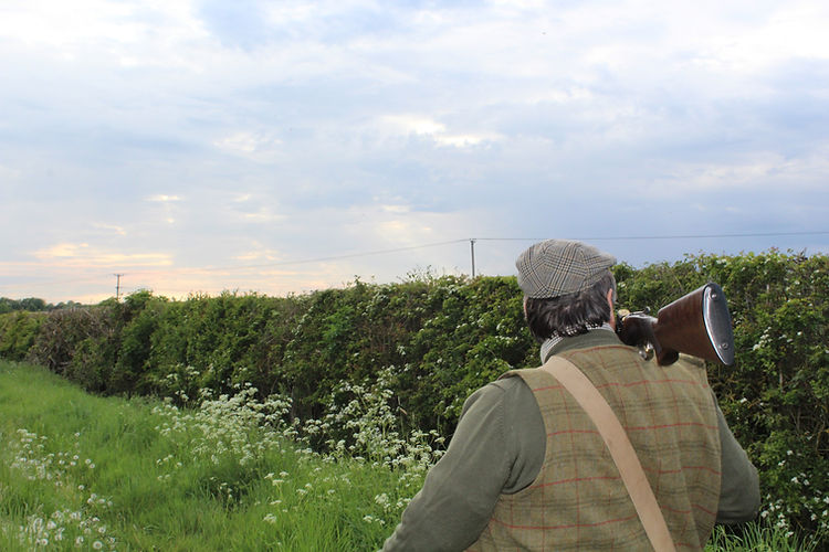 Man with shotgun slung over shoulder stands in rural landscape, facing away from camera along grassy path near bushes under cloudy sky.