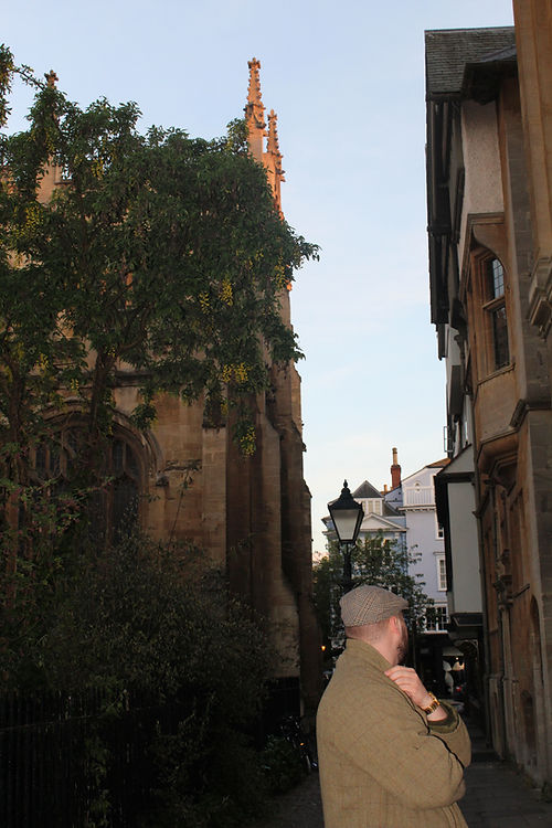 Man in tweed jacket standing on a city street next to a tree and stone buildings, with architecture and street lamp in the background.