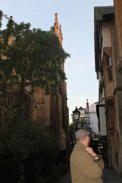 Man in tweed jacket standing on a city street next to a tree and stone buildings, with architecture and street lamp in the background.
