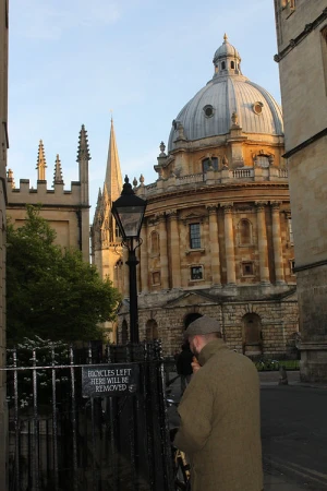 Man in tweed jacket stands in front of Oxford University's Radcliffe Camera building and wrought iron fence on a sunny day.