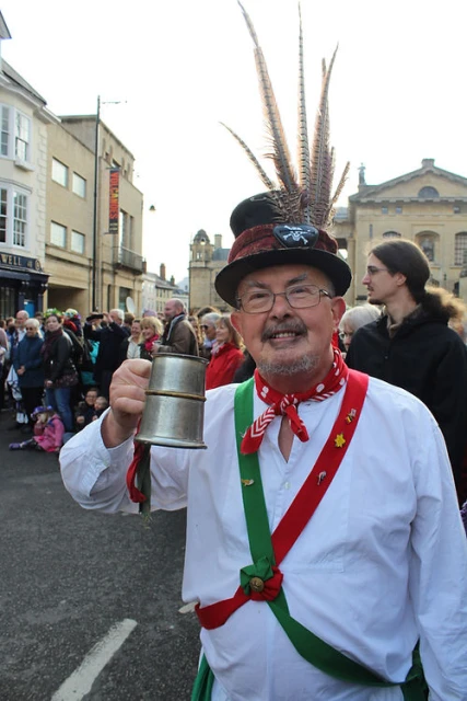 Adult man in traditional English Morris dancing costume holds a metal tankard outdoors in a town square, surrounded by onlookers.