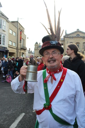 Adult man in traditional English Morris dancing costume holds a metal tankard outdoors in a town square, surrounded by onlookers.