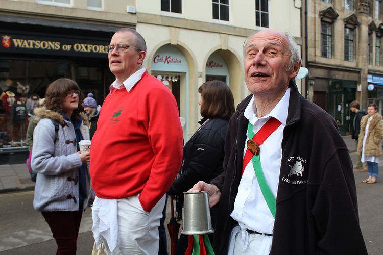 Two mature men stand outside on a city street. The man in the foreground is holding a metal vessel and is wearing a sash.