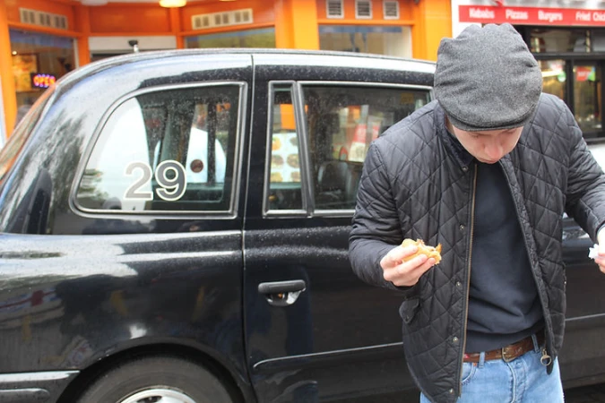 An adult male standing beside a black taxi, looking down at a sandwich in his hands, wearing a quilted jacket, flat cap and jeans. 