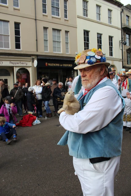 Man in traditional Morris dance attire holding a sheep puppet in a town square surrounded by onlookers and buildings.