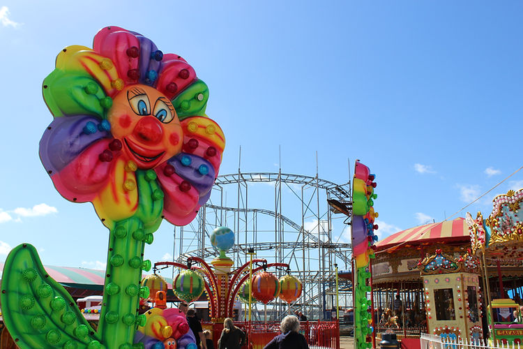 Colorful flower carnival ride in outdoor amusement park setting with blue sky, roller coaster, and vintage carousel in background.