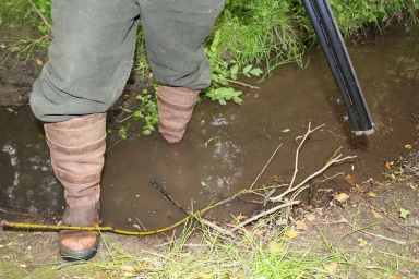 Person wearing green trousers and brown leather wellington boots standing in a shallow muddy stream holding a shotgun