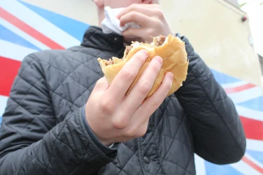 Close-up of a person eating a half-eaten burger while wearing a black quilted jacket with a Union Jack background.
