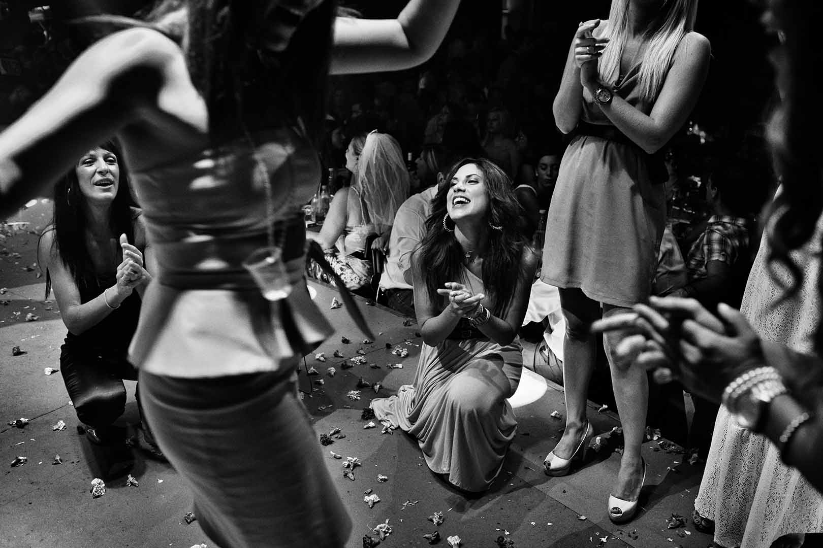 Black and white party photo of women dancing and clapping, surrounded by confetti on the floor, in a dark, indoor environment.