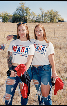 Two young red-haired women pose outdoors in matching MAGA t-shirts with American flags in the background, wearing ripped jeans.
