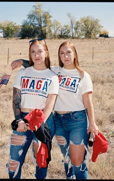 Two young red-haired women pose outdoors in matching MAGA t-shirts with American flags in the background, wearing ripped jeans.