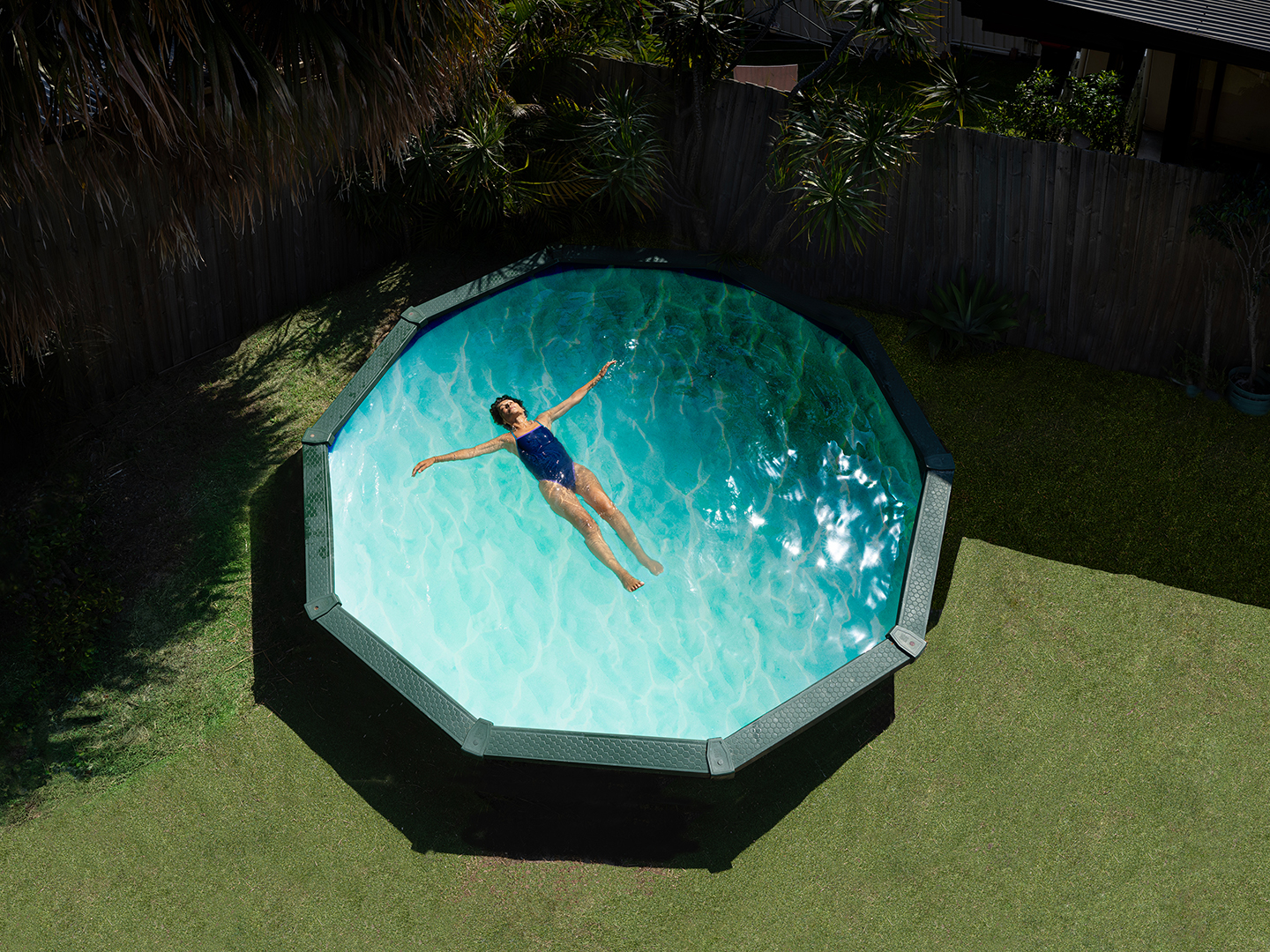 Aerial view of a woman in a blue swimsuit floating in an outdoor above-ground swimming pool in a green backyard on a sunny day.