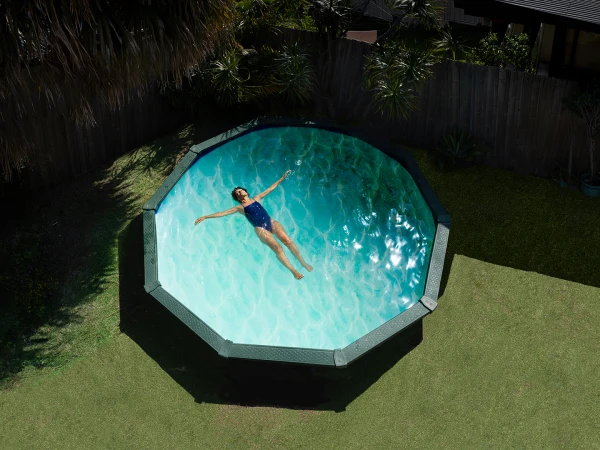 Aerial view of a woman in a blue swimsuit floating in an outdoor above-ground swimming pool in a green backyard on a sunny day.