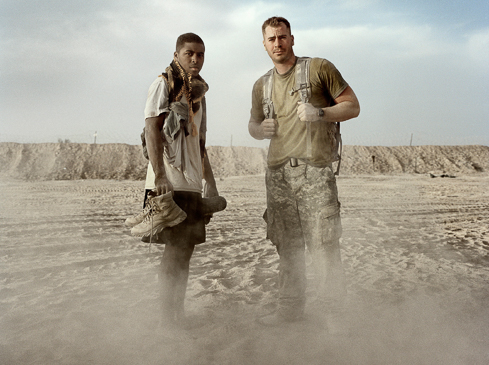 Two male soldiers stand in a desert landscape covered in dust. One carries boots; both wear military attire, conveying resilience and fatigue.