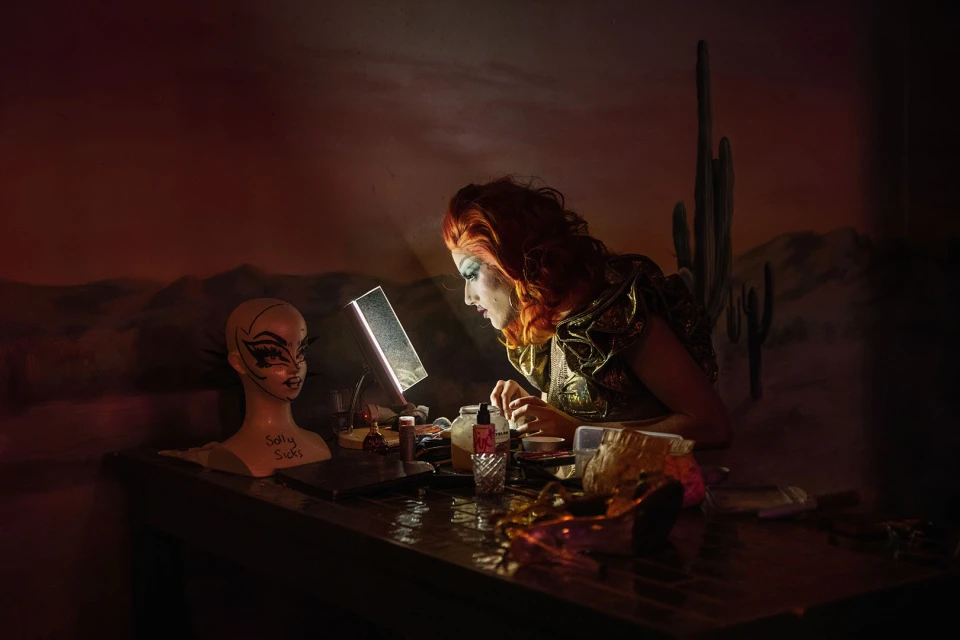 Red-haired drag queen applies stage makeup at a vanity table with a mannequin head, cosmetics, and desert mural backdrop.