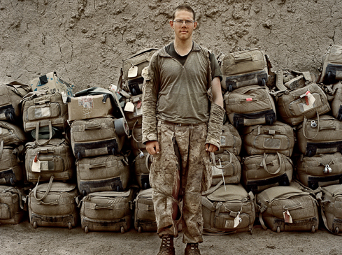 Full shot of a soldier standing in front of a large pile of luggage. The soldier wears camouflage and the background is a neutral color.