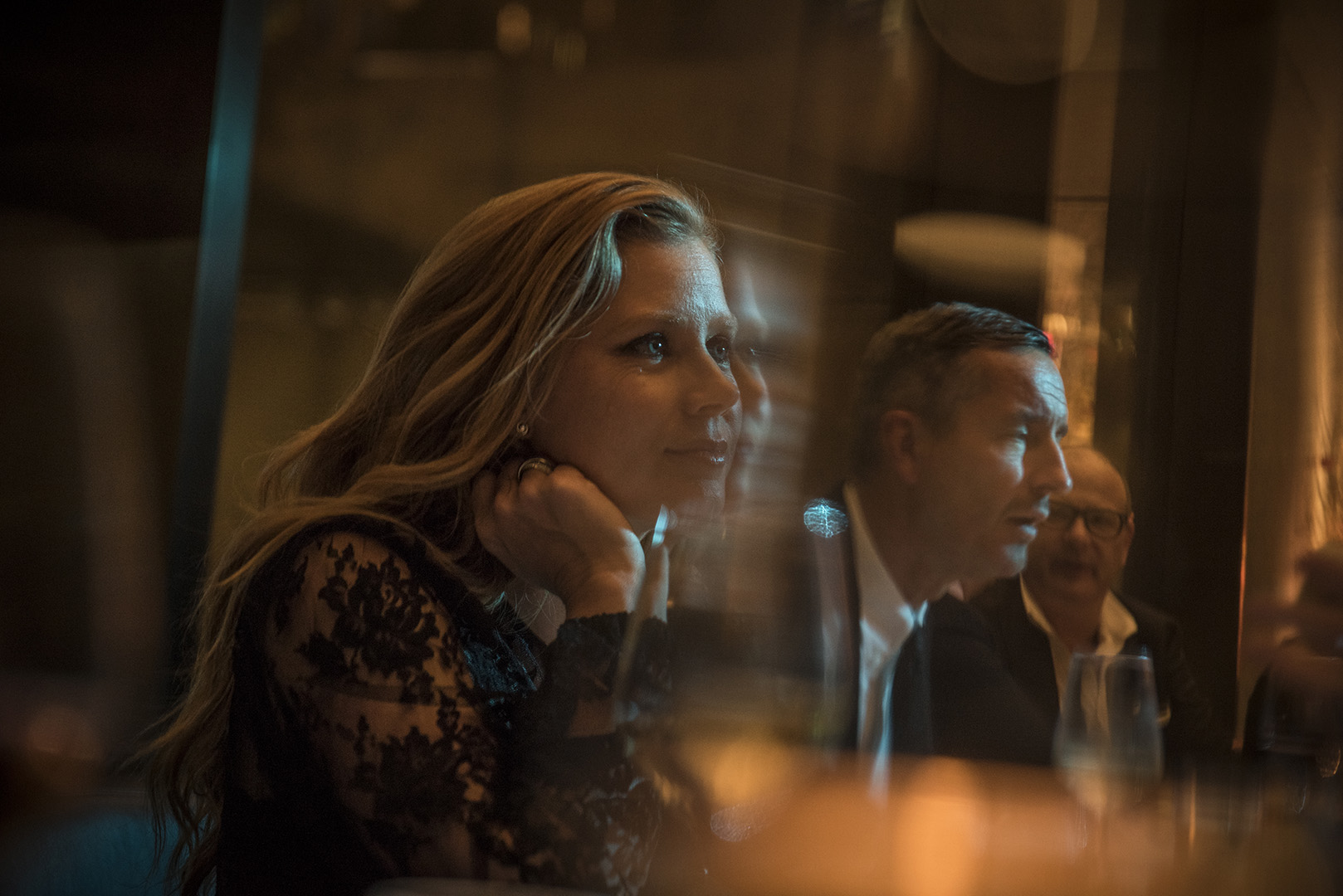 Blonde woman in black lace dress resting her chin on her hand seated at a table with other people by Adam Ferguson