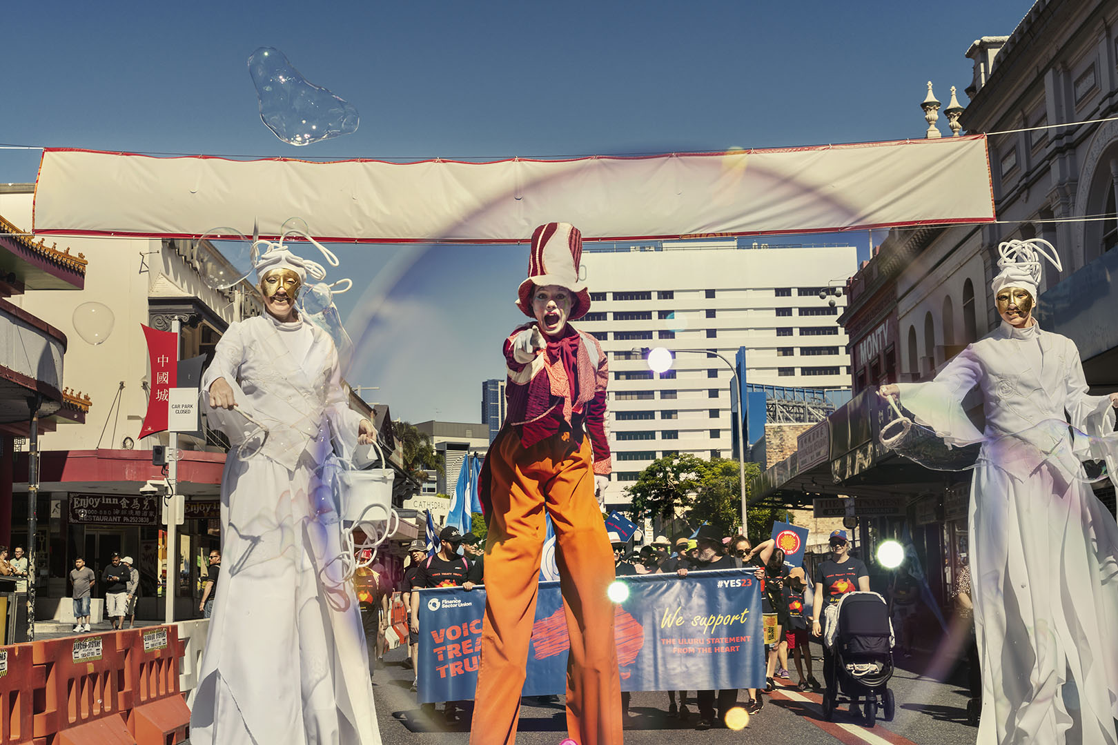 Street parade featuring performers on stilts, costumes, and a large banner, with a city building in the background on a sunny day.