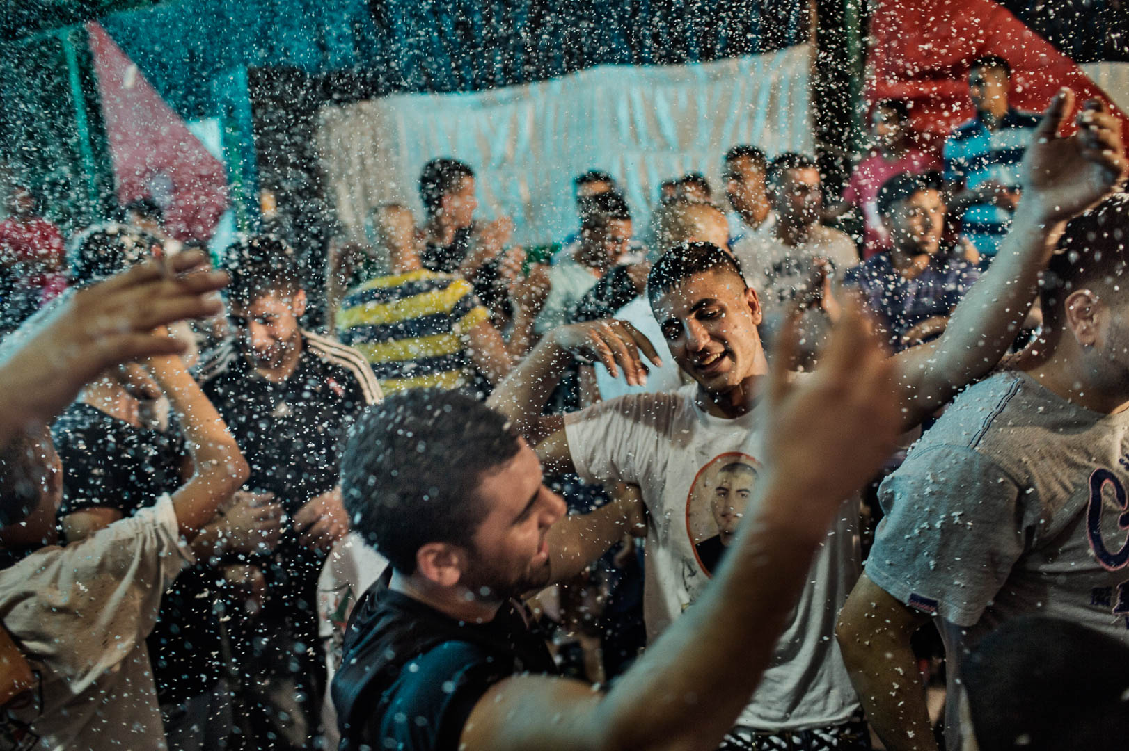 Large group of young adults celebrate indoors during an event with artificial snow, wearing casual clothing and raising their arms.