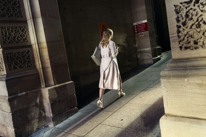 A woman in a coat walks under an archway with columns while looking at her phone. The scene is illuminated by directional lighting.