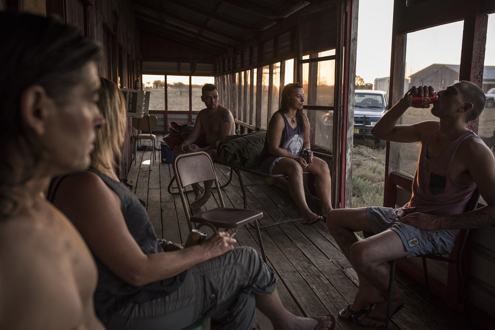 Several adults relax on a porch in an outdoor setting, one drinking from a can, creating an intimate, casual gathering at dusk.