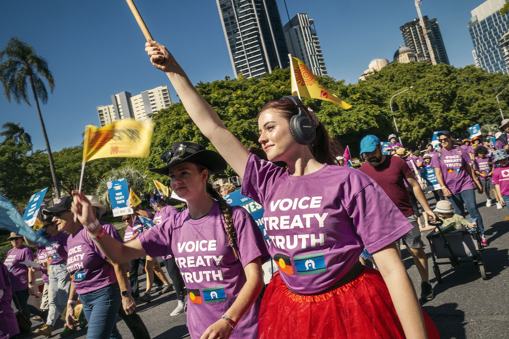 Crowd marches for Indigenous rights, holding signs and flags. Women wear purple "Voice Treaty Truth" shirts in sunny urban setting.