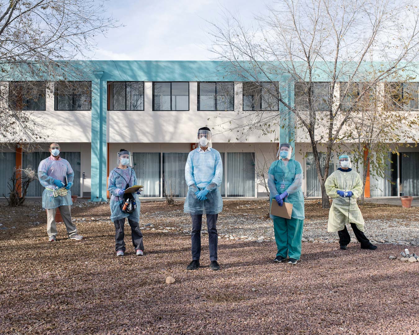 A group of healthcare workers in personal protective equipment stand outside of a building with a gravel surface in the foreground.